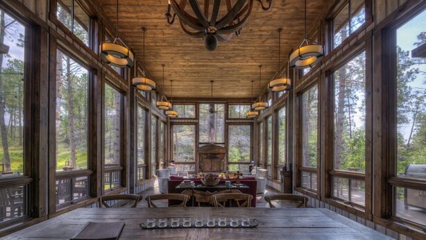 A wooden dining area with a long table and chairs, large windows, and hanging light fixtures. - Majestic Cabin near Mount Rushmore (Keystone)