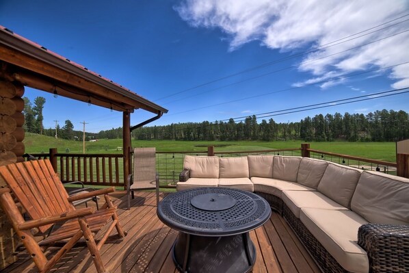 A wooden deck with a curved sofa and a round table, overlooking a green field and trees. - Cabin close to Nemo, private stocked pond and hot tub great for families! (Nemo)