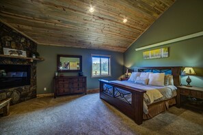 A bedroom with a stone fireplace, a large bed, a wooden ceiling, and a window with a view.
