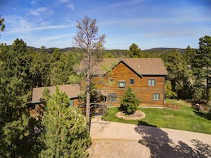 A two-story log cabin with a front porch, surrounded by a well-maintained lawn and dense forest.