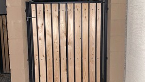 Une porte en bois, agrémentée de barreaux en métal noir, est installée dans un espace recouvert de gravier, avec un mur blanc en arrière-plan.