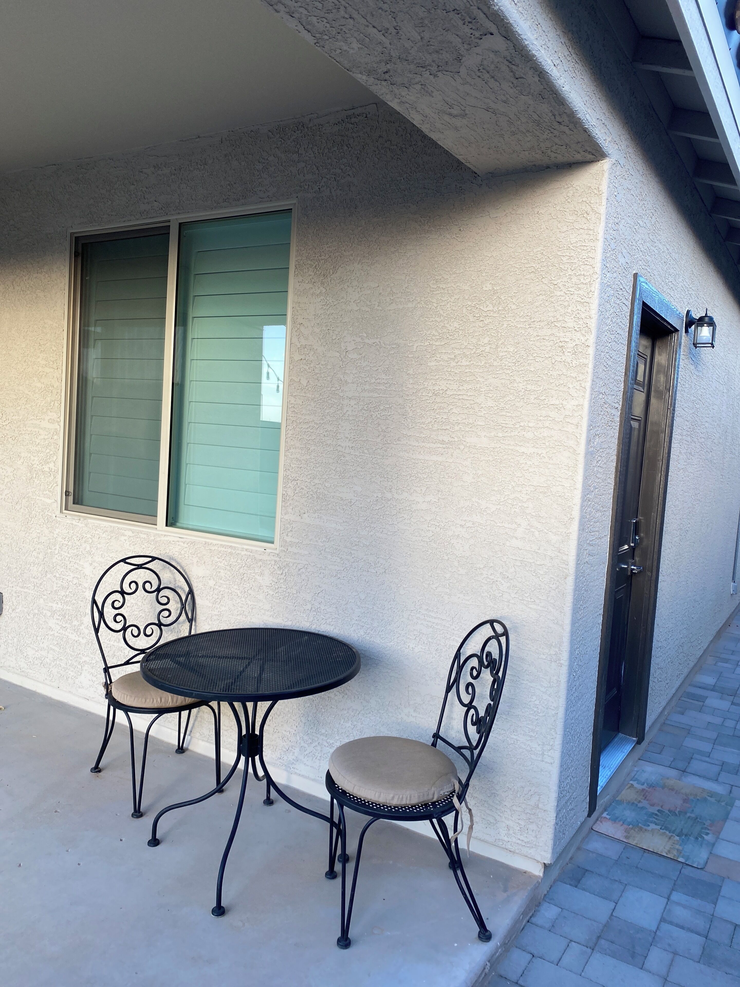 A patio with a round table and two chairs, a window with shutters, and a door with a light fixture.