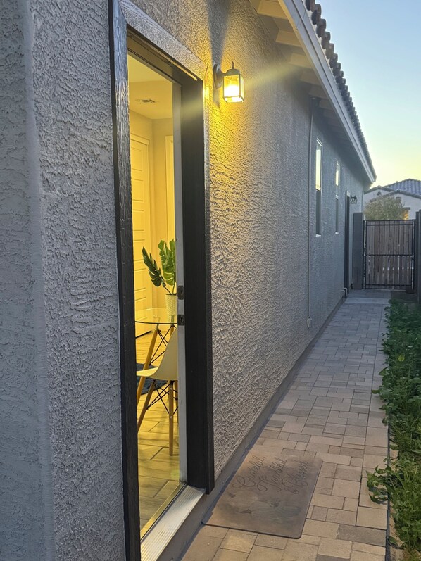A house with a textured exterior, a door leading to a room with a chair and a potted plant, and a pathway with a welcome mat.