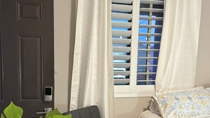Une pièce avec une porte sombre, un fauteuil gris, une fenêtre avec des stores, un lit recouvert d’une couvre-lit à motifs et une grande plante verte.