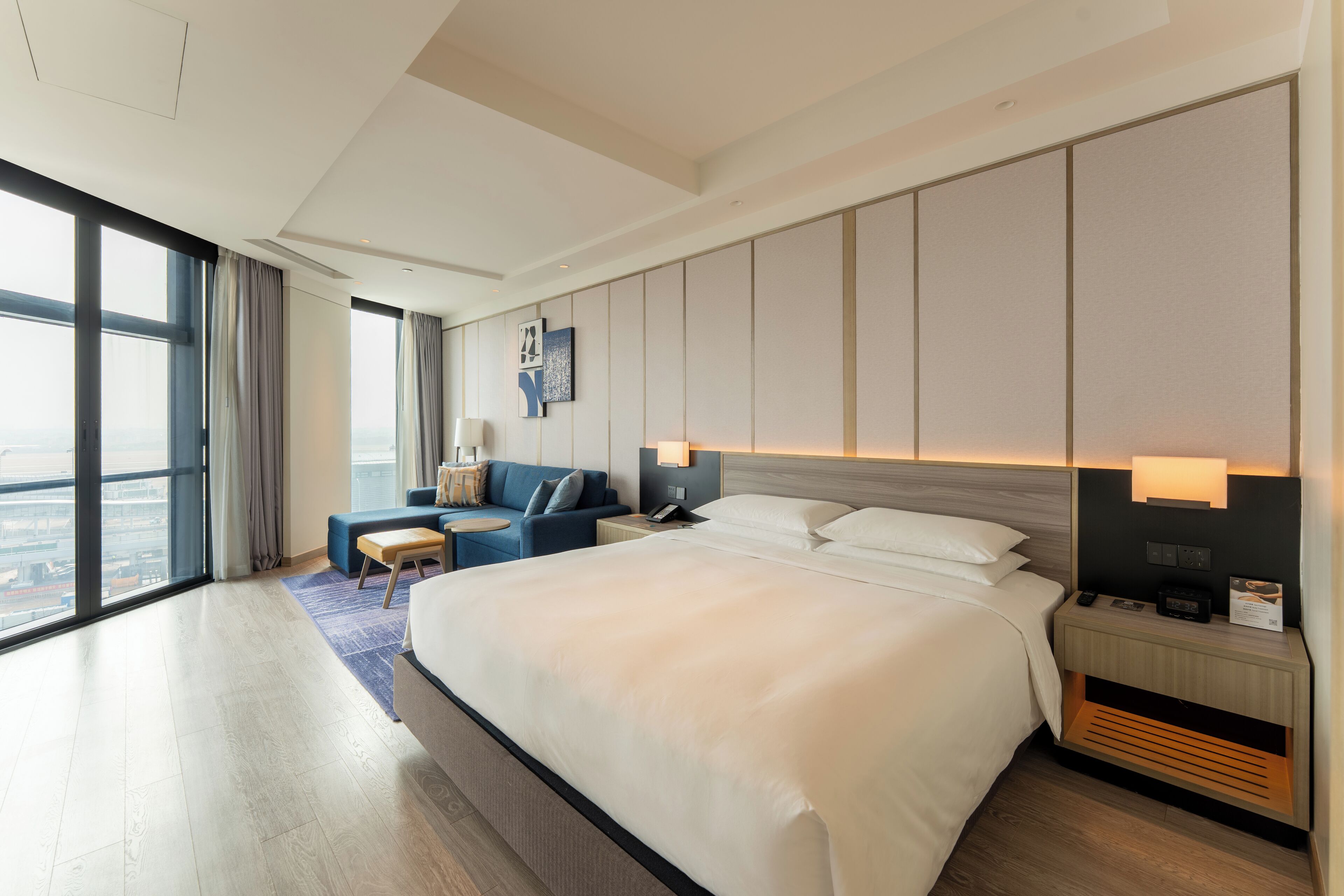 A modern hotel room with a large bed, a seating area with a sofa and armchair, a desk with a lamp, and a city view through large windows.