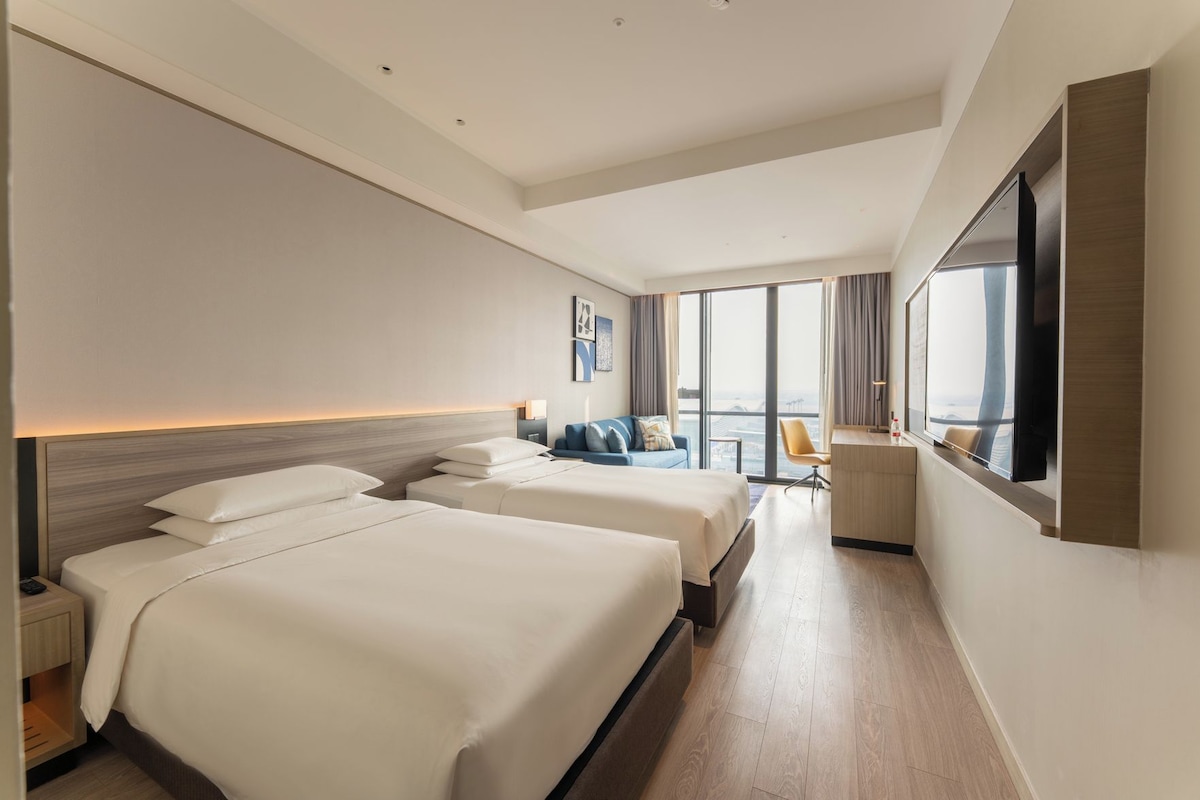 A hotel room with two beds, a desk, a chair, and a large window offering a city view.