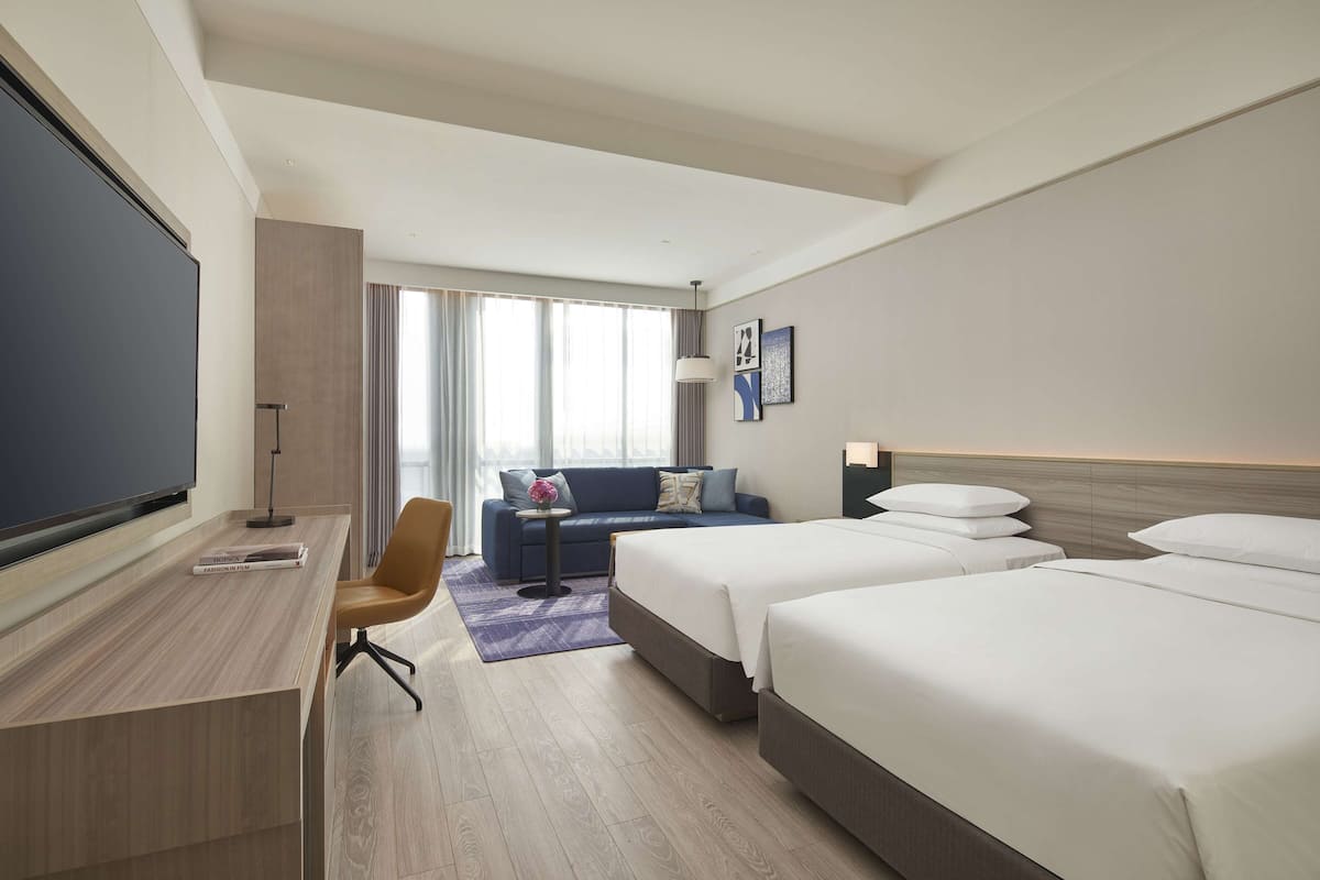 A hotel room with a large flat-screen TV, a desk with a chair, a sofa, and two beds.