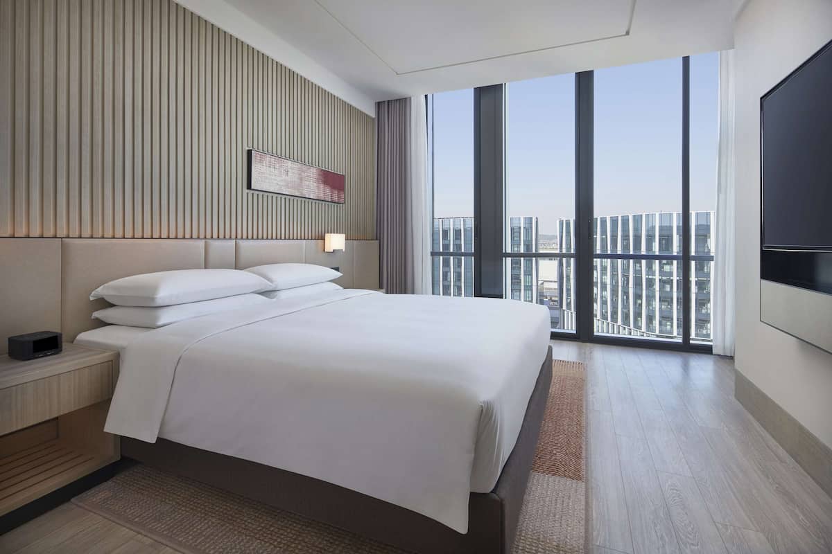 A modern hotel room with a large bed, a flat-screen TV, and a city view through large windows.