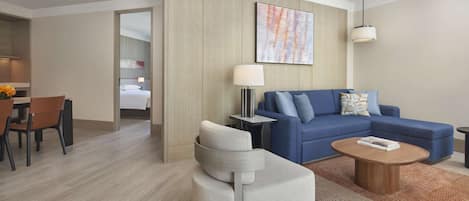 A modern hotel room with a blue sofa, a white armchair, a wooden coffee table, and a dining area with a table and chairs.
