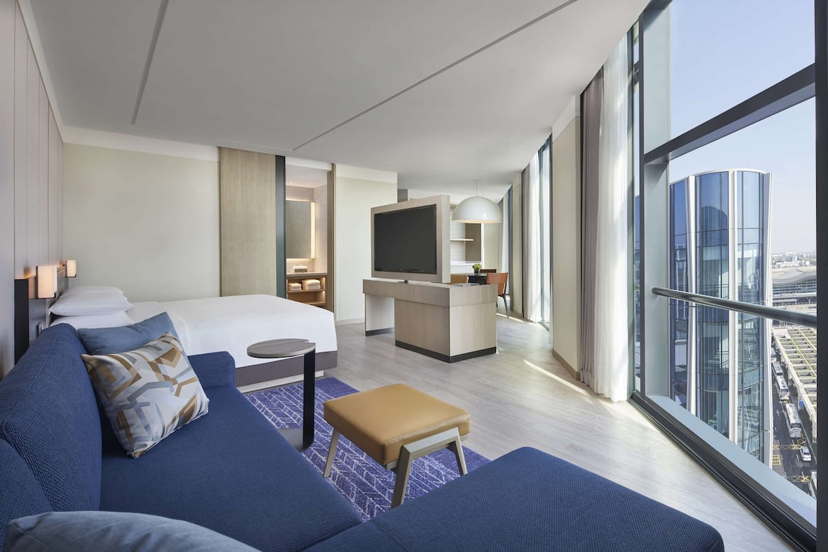 A modern hotel room with a large bed, a sofa, a desk with a TV, and a city view.