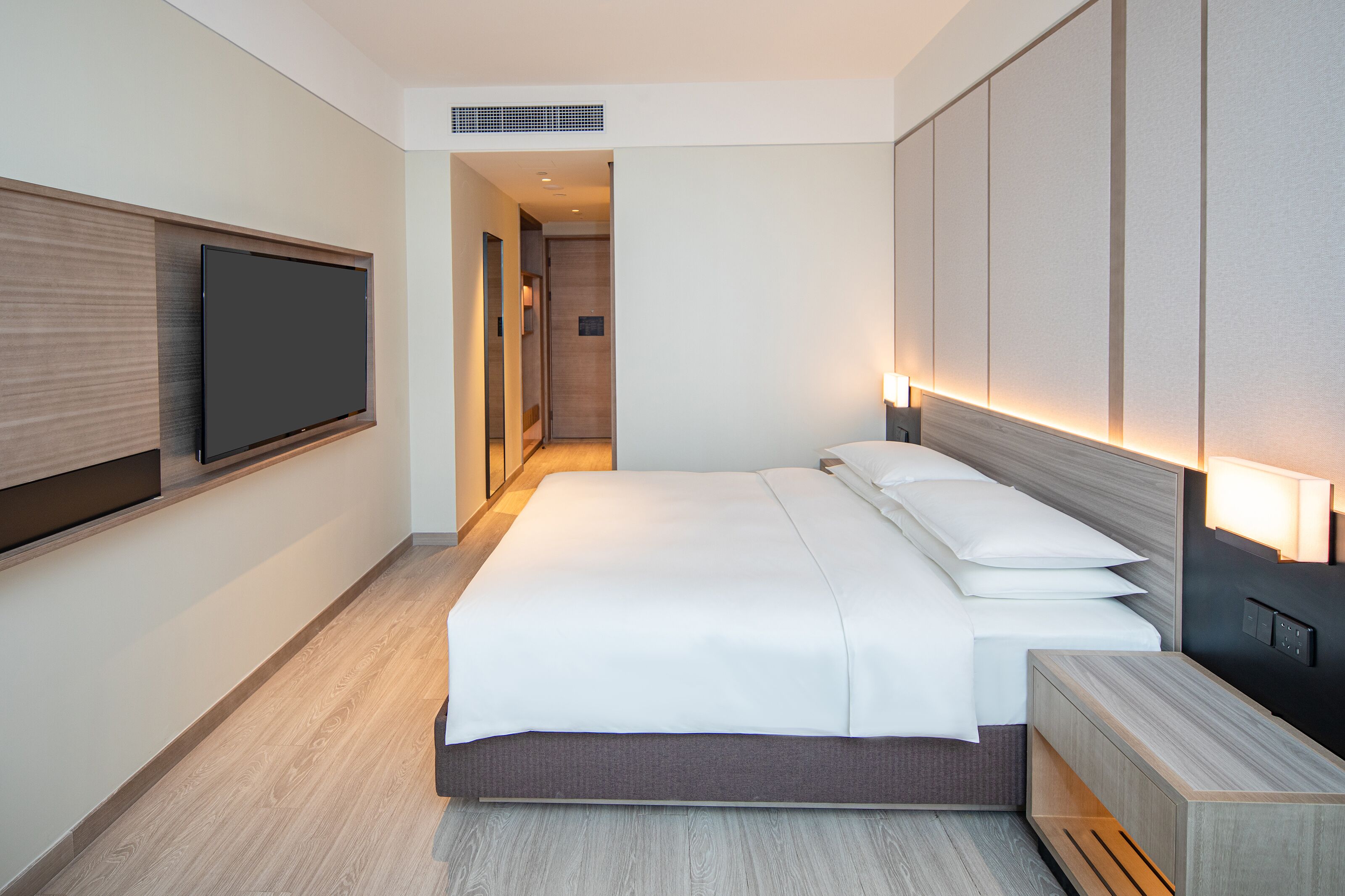 Una habitación de hotel moderna con una cama grande, un televisor de pantalla plana montado en la pared y una mesita de noche con una lámpara.