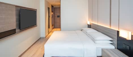 A modern hotel room with a large bed, a flat-screen TV mounted on the wall, and a bedside table with a lamp.