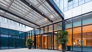 Een moderne entree van een gebouw met glazen deuren, een overdekte doorgang en twee potplanten.