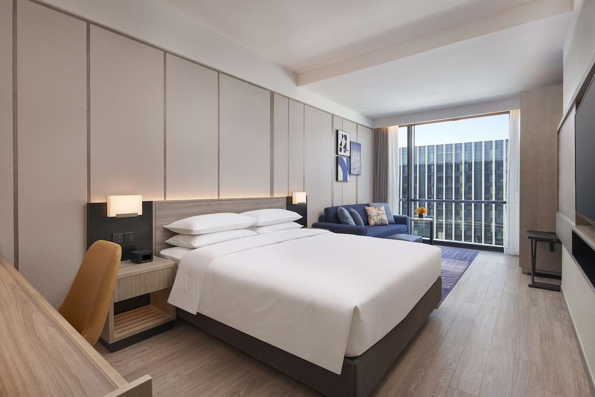 A modern hotel room with a large bed, a desk, a sofa, and a balcony with a city view.