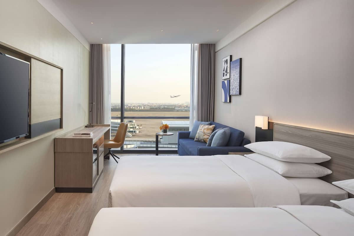 A hotel room with a large window, a bed, a desk, a chair, and a small table with a flower vase.