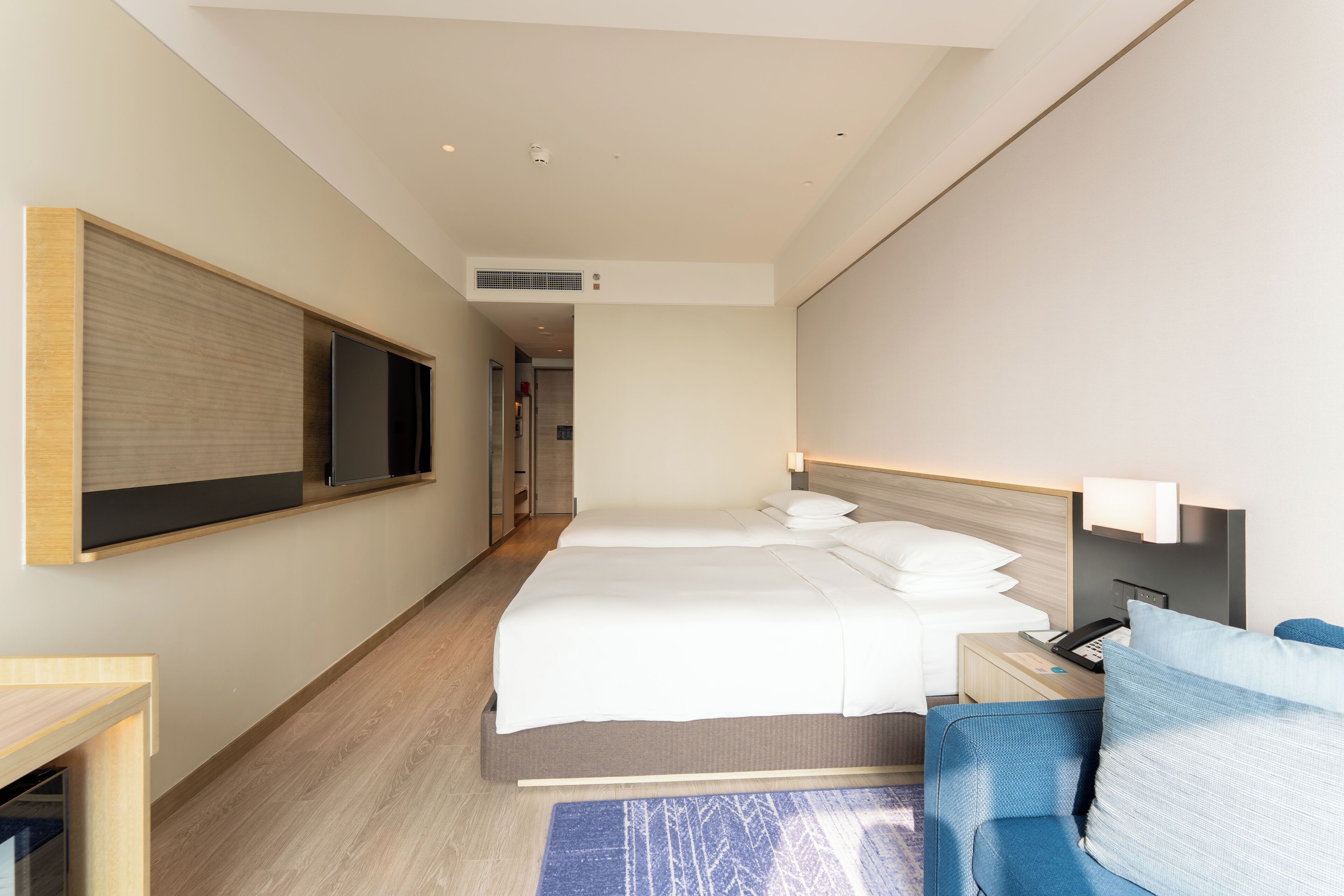 A modern hotel room with a large bed, a TV mounted on the wall, a desk with a lamp, and a blue sofa.
