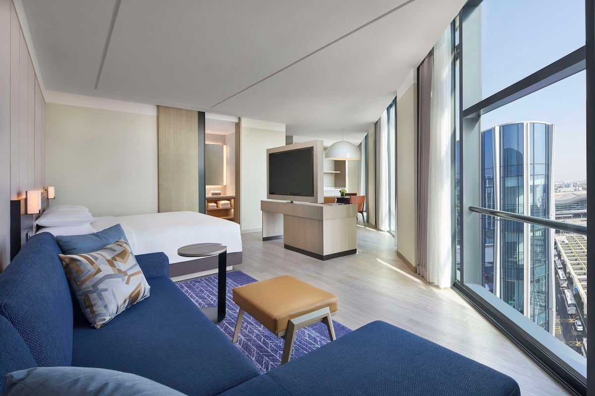 A modern hotel room with a bed, sofa, TV, desk, and large windows with a city view.