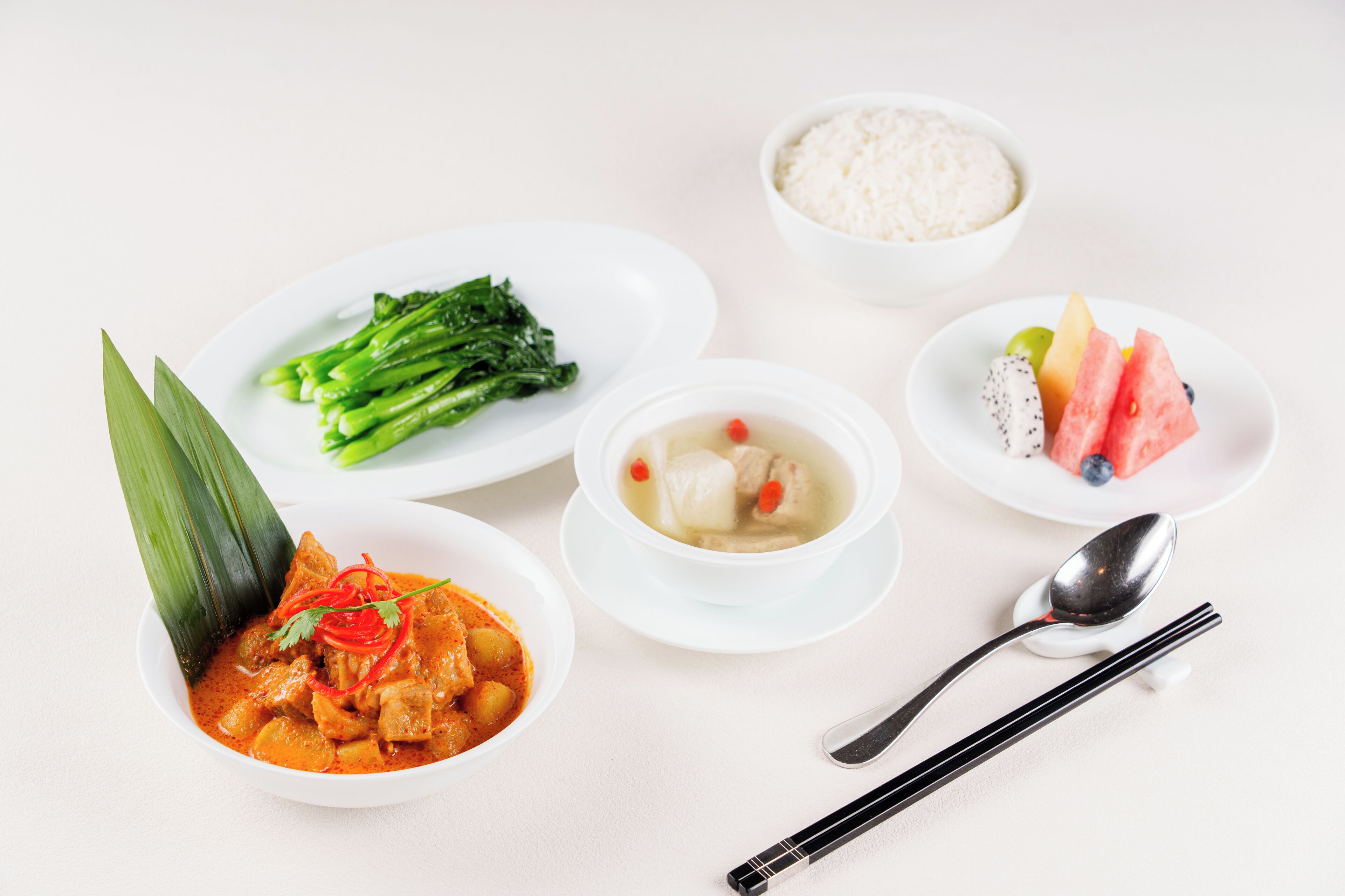 A table with various dishes including a bowl of soup, a plate of steamed vegetables, a bowl of rice, and a plate with watermelon and other fruits.