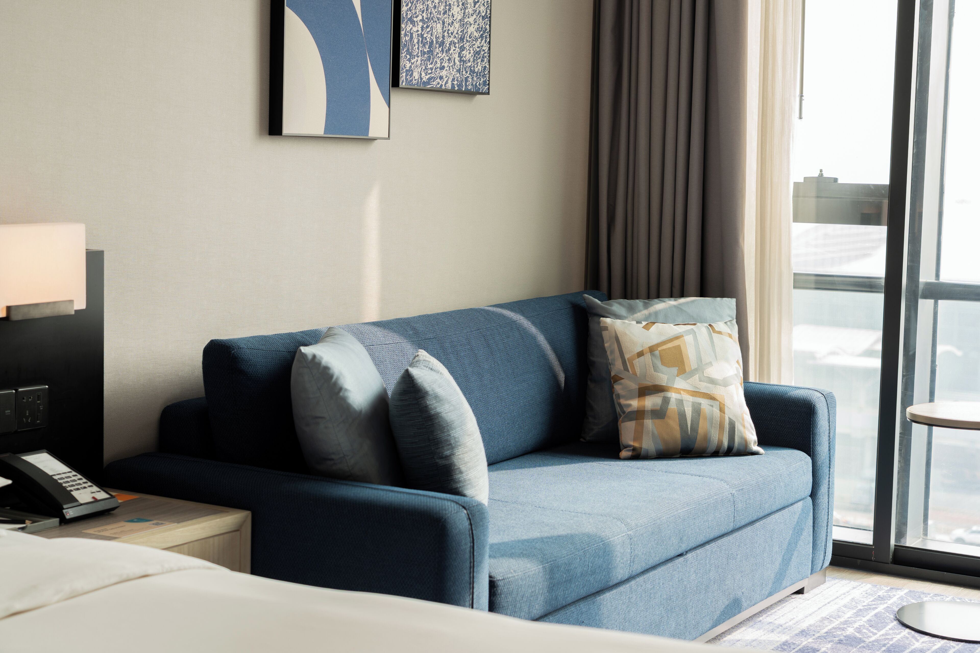 A modern hotel room with a blue sofa, a bedside table with a lamp, and a window with a view of the city.