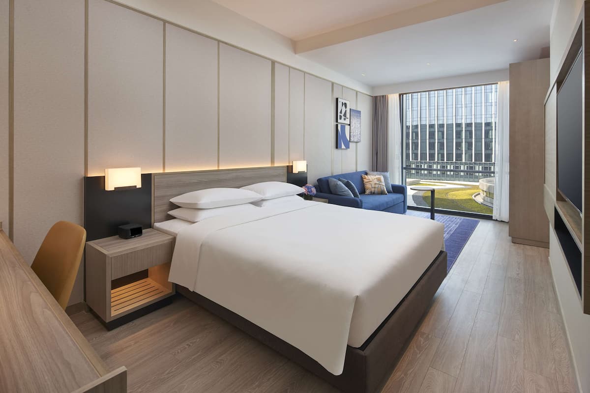 A modern hotel room with a large bed, a desk, a chair, and a sofa. There is a large window with a view of the city.