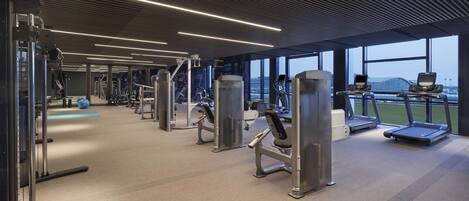 A modern gym with various exercise equipment, large windows, and a view of the cityscape.