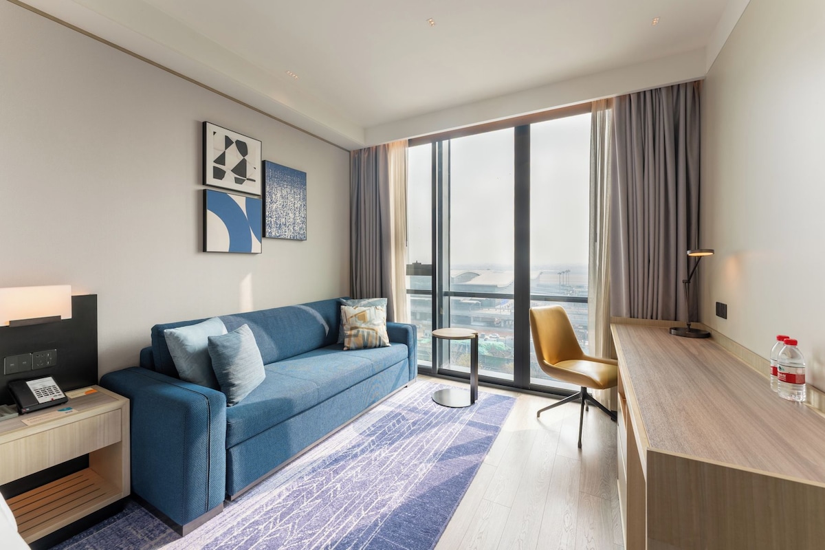 A modern hotel room with a blue sofa, a small desk, a chair, a nightstand, and a large window offering a city view.