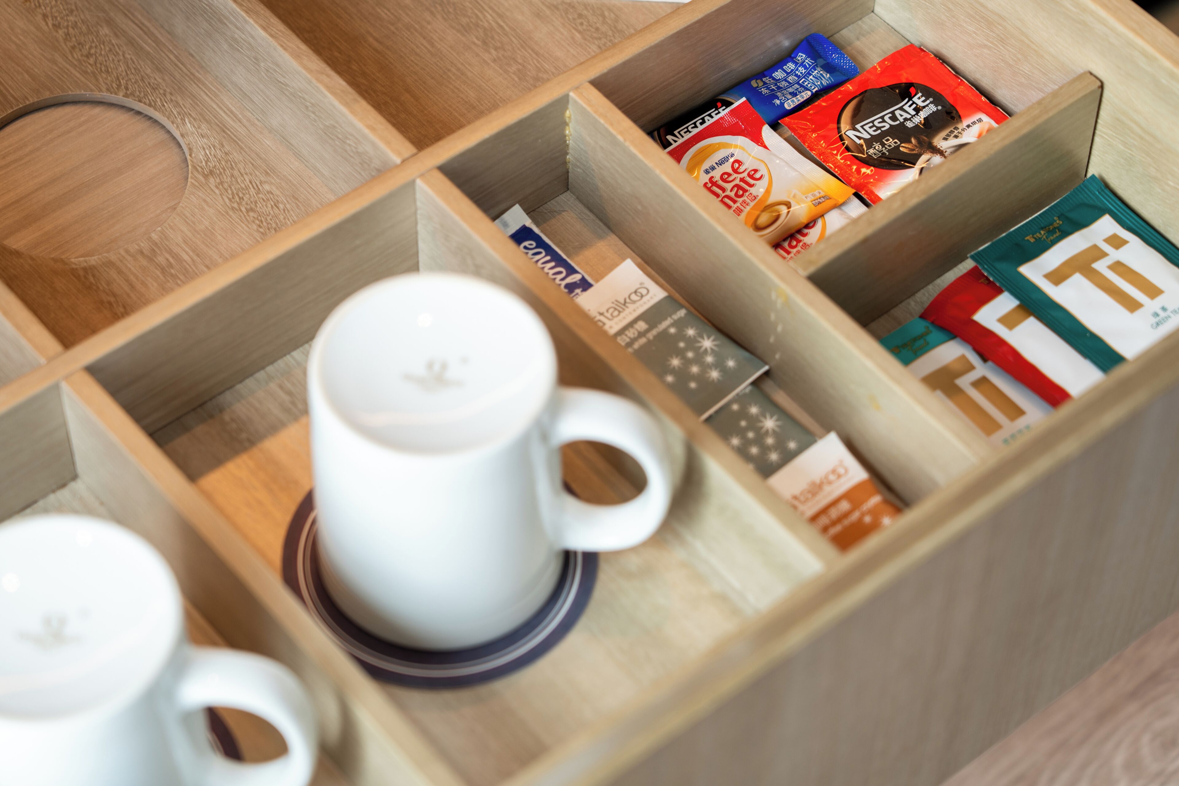 A wooden coffee station with a white coffee maker and cups, and a box of Nespresso capsules.