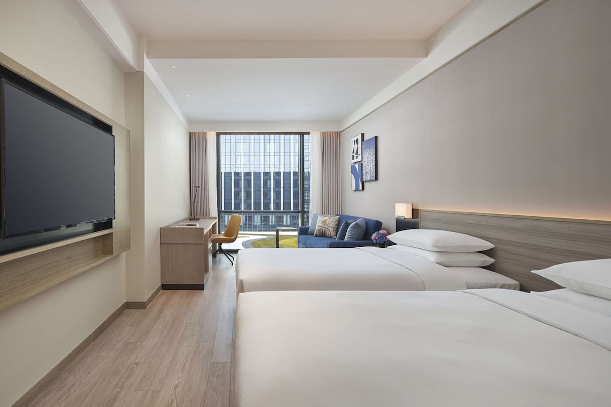 A hotel room with a large bed, a smaller bed, a desk, a chair, a TV, and a window with a view of the city.