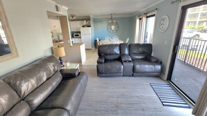 Un salon avec un canapé en cuir noir, une table à manger avec des chaises, un coin cuisine et une vue à travers une porte-fenêtre coulissante.