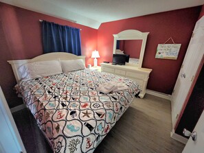 Quarto com cama, mesa de cabeceira, abajur, espelho e um aviso na parede.