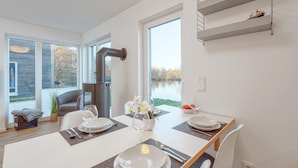 Un espace repas moderne avec une table dressée pour deux personnes, une grande fenêtre donnant sur un lac et un poêle à bois encastré.