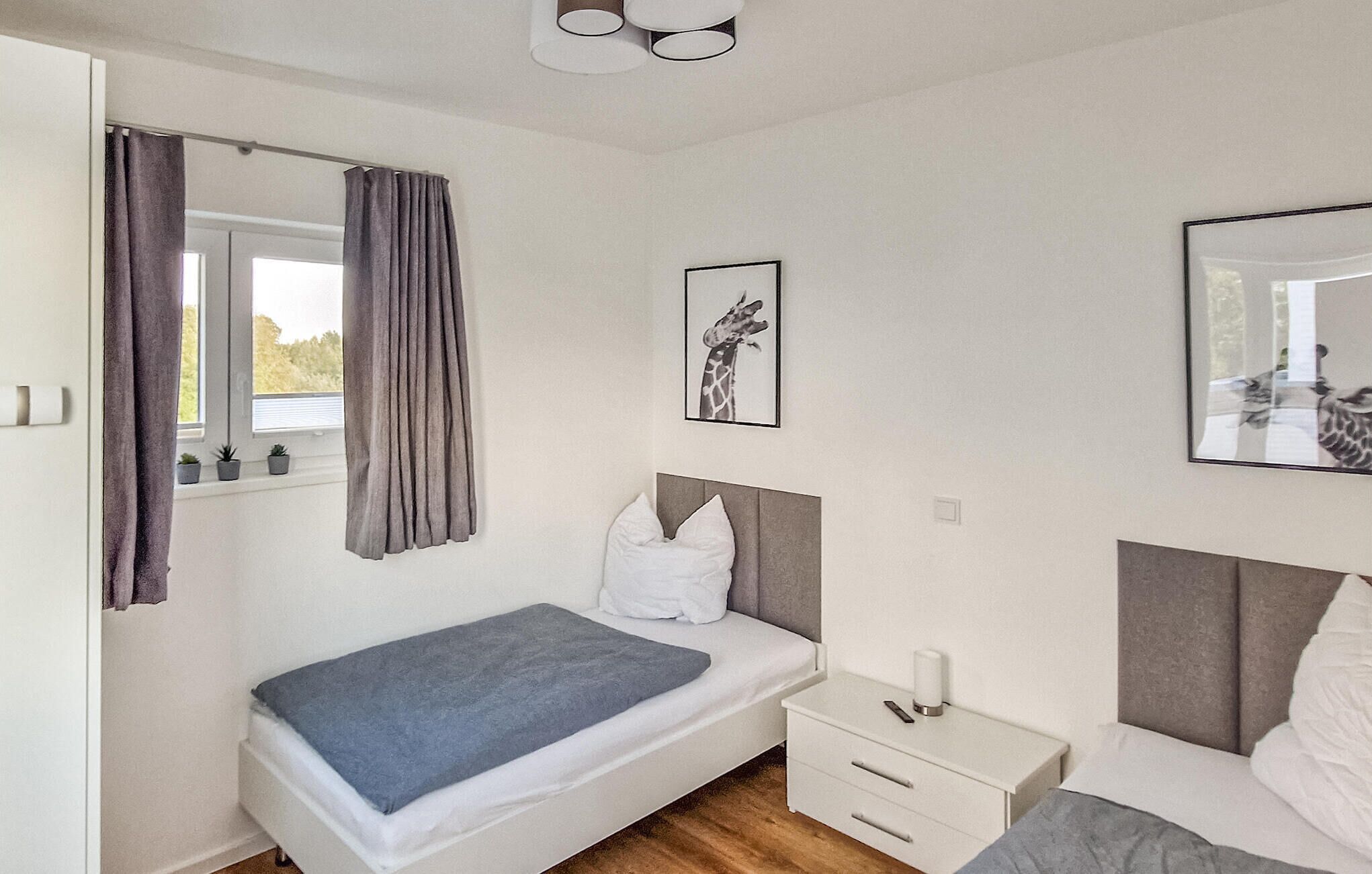 A bedroom with a bed, bedside table, and a window with curtains. There are two framed pictures on the wall.