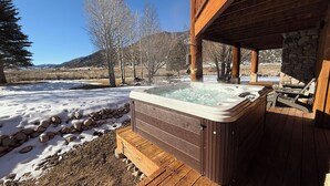 Een hottub op een houten terras, met een stenen muur en houten balken, omgeven door een besneeerd landschap en bomen.