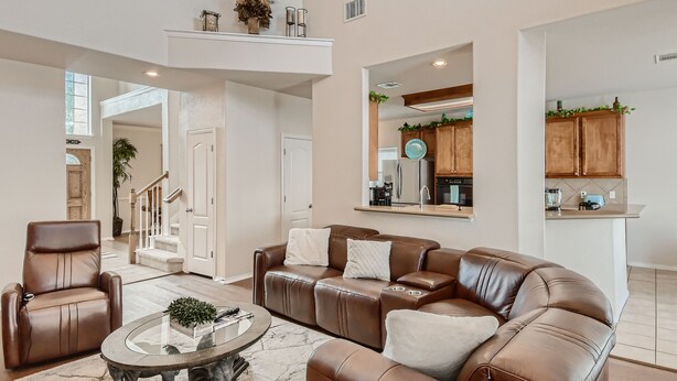 A spacious living room with a brown leather sofa, a glass coffee table, and a brown leather armchair. A staircase leads to an upper level, and a kitchen is visible in the background. - Home with private pool rents for longer term stay (Cedar Park)