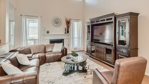 A spacious living room with a brown leather sectional sofa, a glass coffee table with elephant legs, a fireplace, and a large dark wooden cabinet. - Home with private pool rents for longer term stay (Cedar Park)