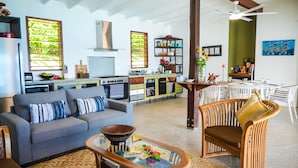 Ein geräumiger Wohnbereich mit einer Couch, einem Couchtisch und Korbstühlen. Im Hintergrund ist eine Küche mit Essbereich und Stühlen zu sehen.