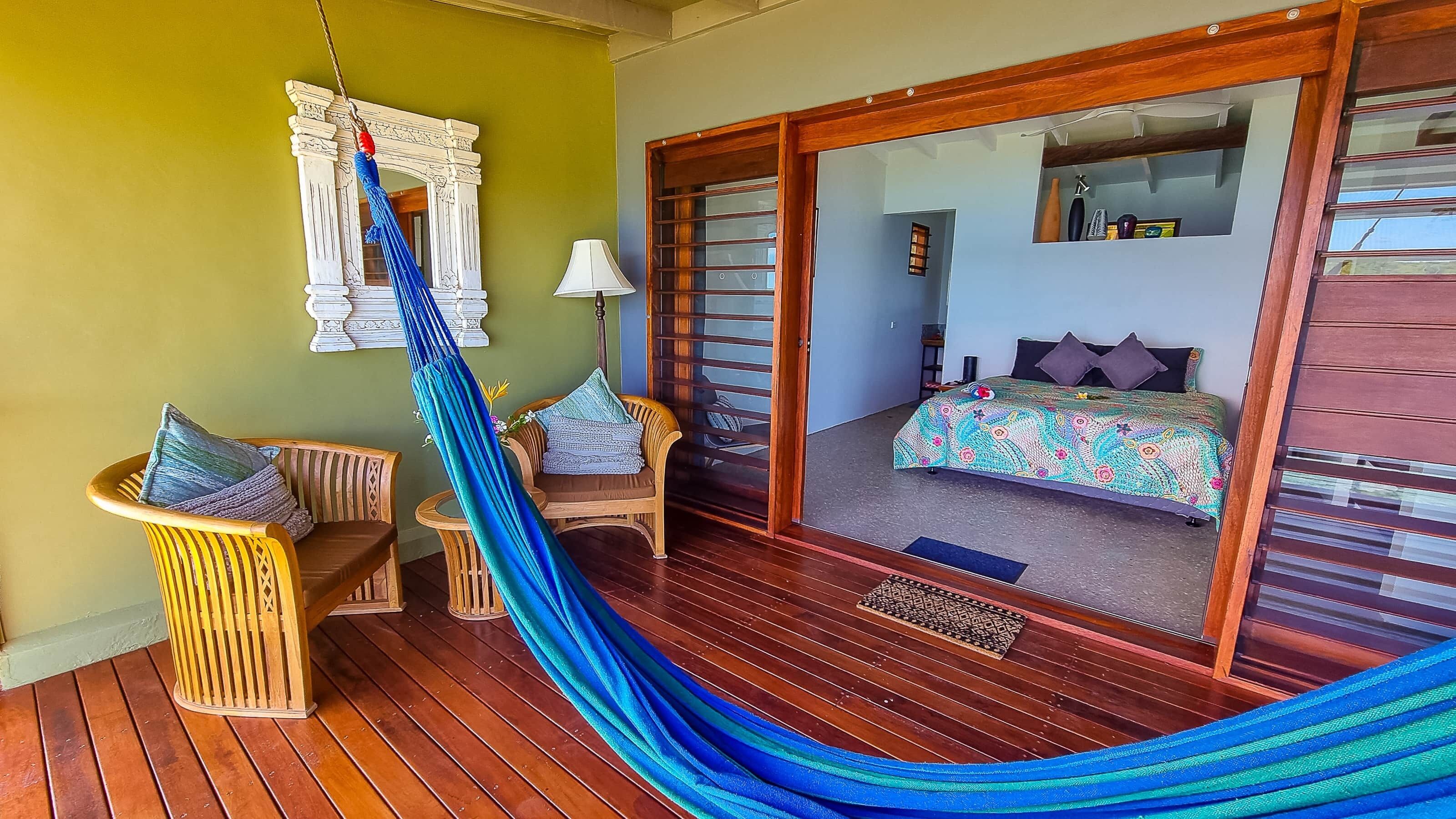 Una habitación con suelo de madera, una hamaca, muebles de mimbre, una cama con colcha colorida y un espejo con marco de madera.