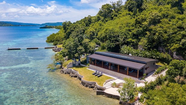 Ein modernes Haus mit einer großen Terrasse mit Blick auf einen klaren, blauen See, umgeben von üppiger Vegetation.