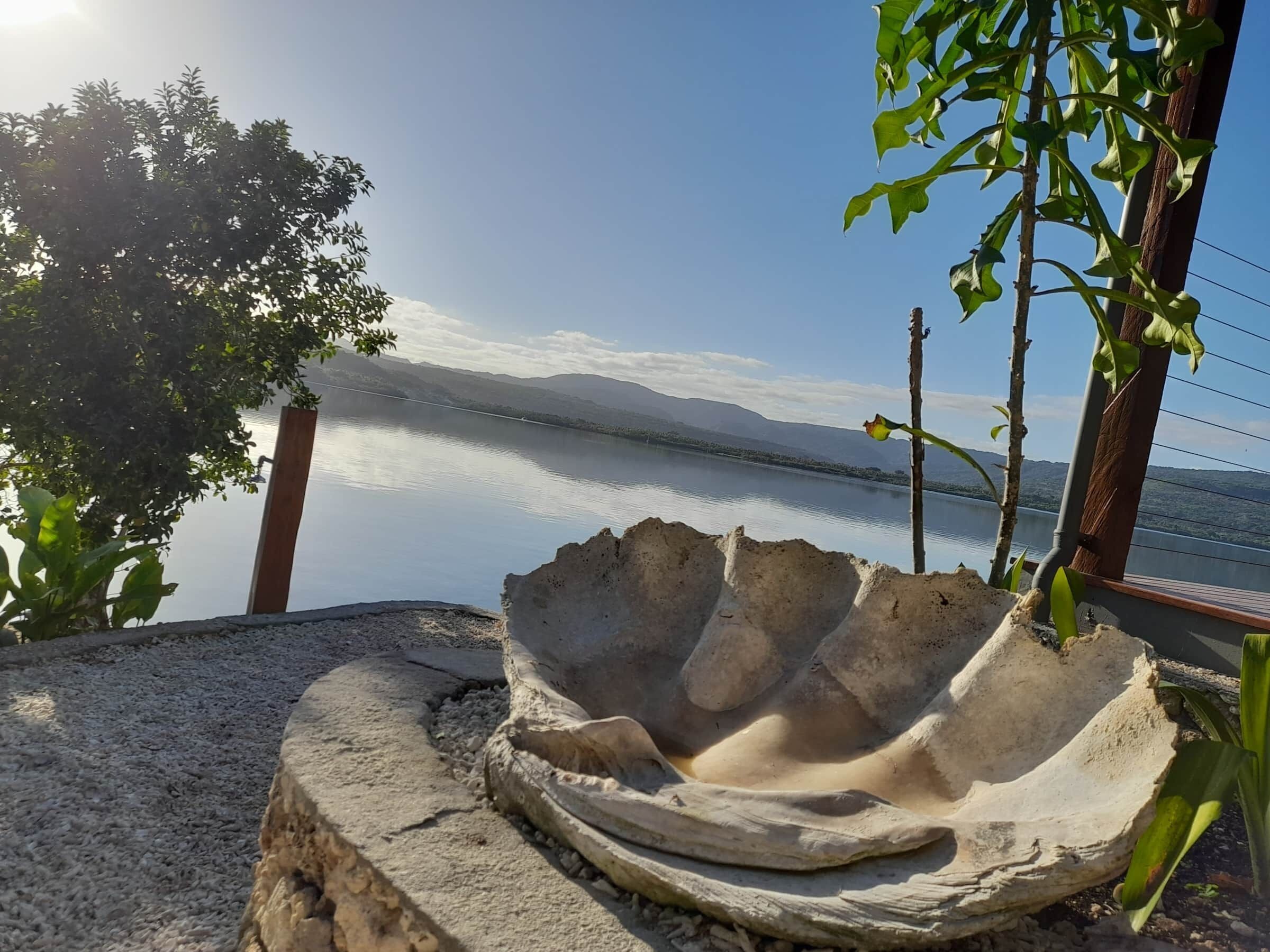 Un bassin en béton aux bords incurvés, entouré de gravier, offrant une vue sur un lac paisible et des montagnes lointaines sous un ciel dégagé.