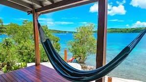 Eine Hängematte auf einer Holzterrasse mit Blick auf das klare, blaue Meer, ein Boot in der Ferne und üppiges Grün.