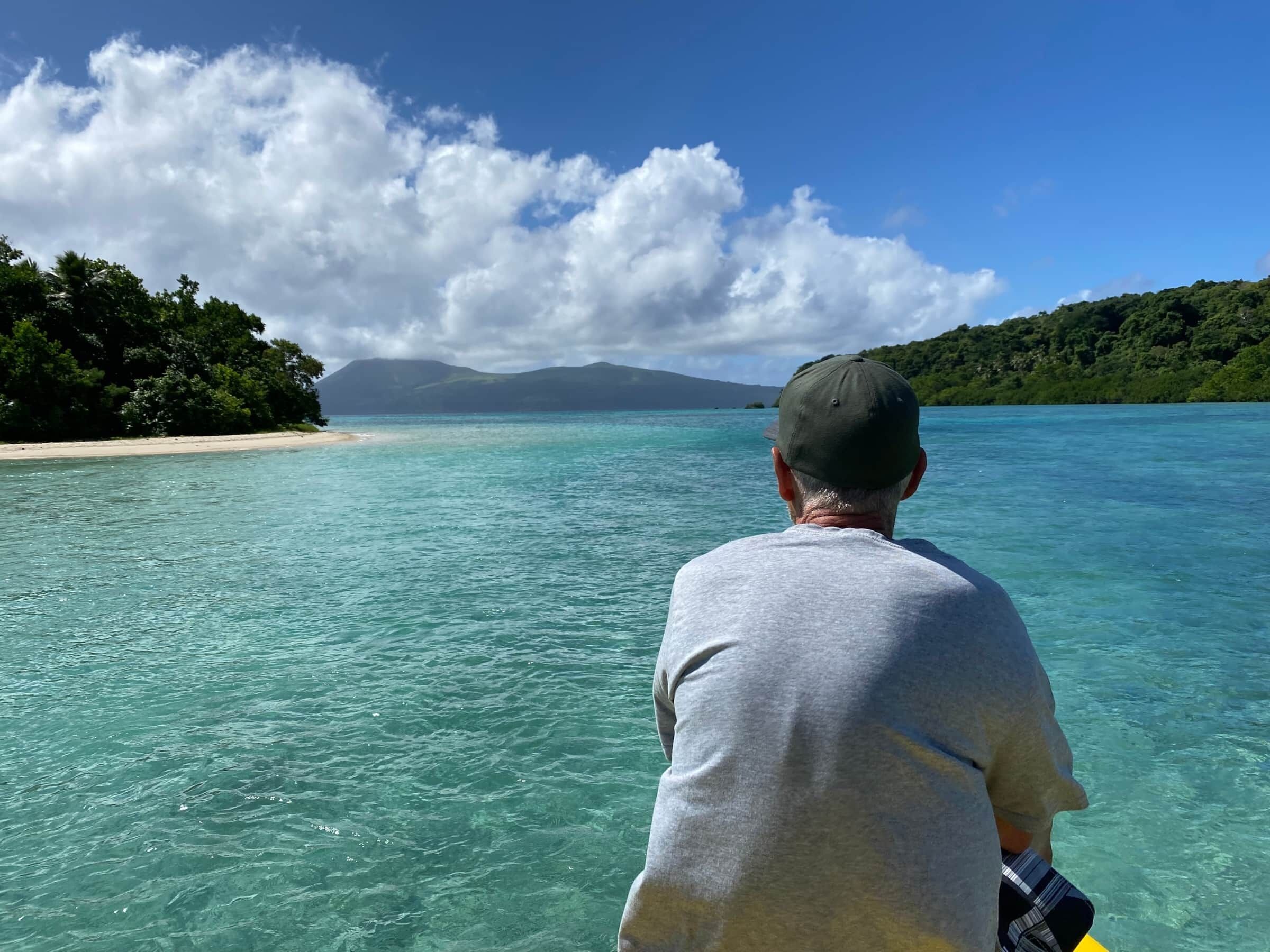 Un uomo con una camicia e un cappello grigi, di fronte a un mare turchese limpido con una spiaggia e colline ricoperte di alberi sullo sfondo.