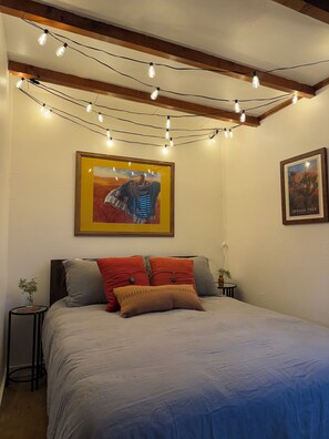 Ein Schlafzimmer mit einem Bett, Kissen, einem gerahmten Bild, Lichterketten und einem kleinen Tisch mit einer Pflanze.