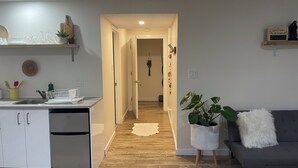 A modern kitchen with white cabinets, a stainless steel refrigerator, and a wooden countertop. There is a hallway leading to a living area with a gray sofa and a white fluffy pillow. - The Tranquil sun room (St. Catharines)