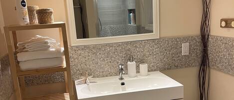 A bathroom with a white sink, a mirror above it, a wooden shelf with towels, and a potted plant.