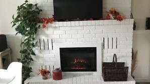 A white brick fireplace with a flat-screen TV mounted above it, surrounded by a white mantelpiece. - The Tranquil sun room (St. Catharines)