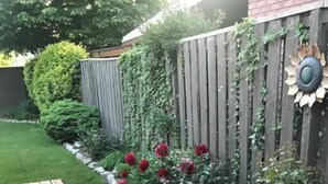 A wooden deck with a stone border and a grassy area with red flowers and green shrubs. - The Tranquil sun room (St. Catharines)