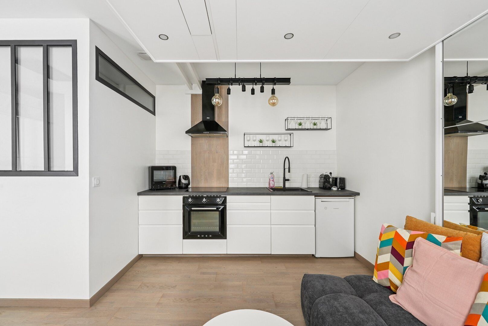 Une cuisine moderne avec des meubles blancs, une cuisinière noire et une machine à café. Le sol est en bois et il y a un canapé gris agrémenté de coussins colorés.