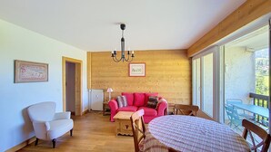 A room with a wooden ceiling, a chandelier, a red sofa, a white armchair, a small table with a plaid tablecloth, and a view through large windows. - 1 RC Gentianes - (Megève)