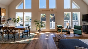 Una habitación amplia con techo alto, ventanas grandes, zona de comedor y sala de estar con televisor.