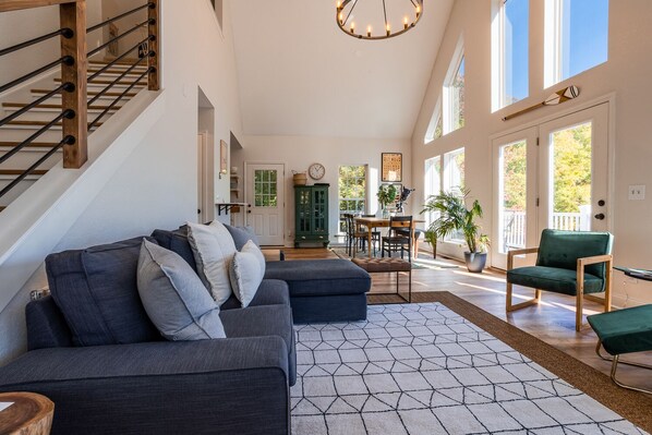 A spacious living area with a dark grey sectional sofa, a green armchair, and a dining table with chairs. - Sunrise Cove | Lake Fontana with Private Dock and Lake Views (Almond)